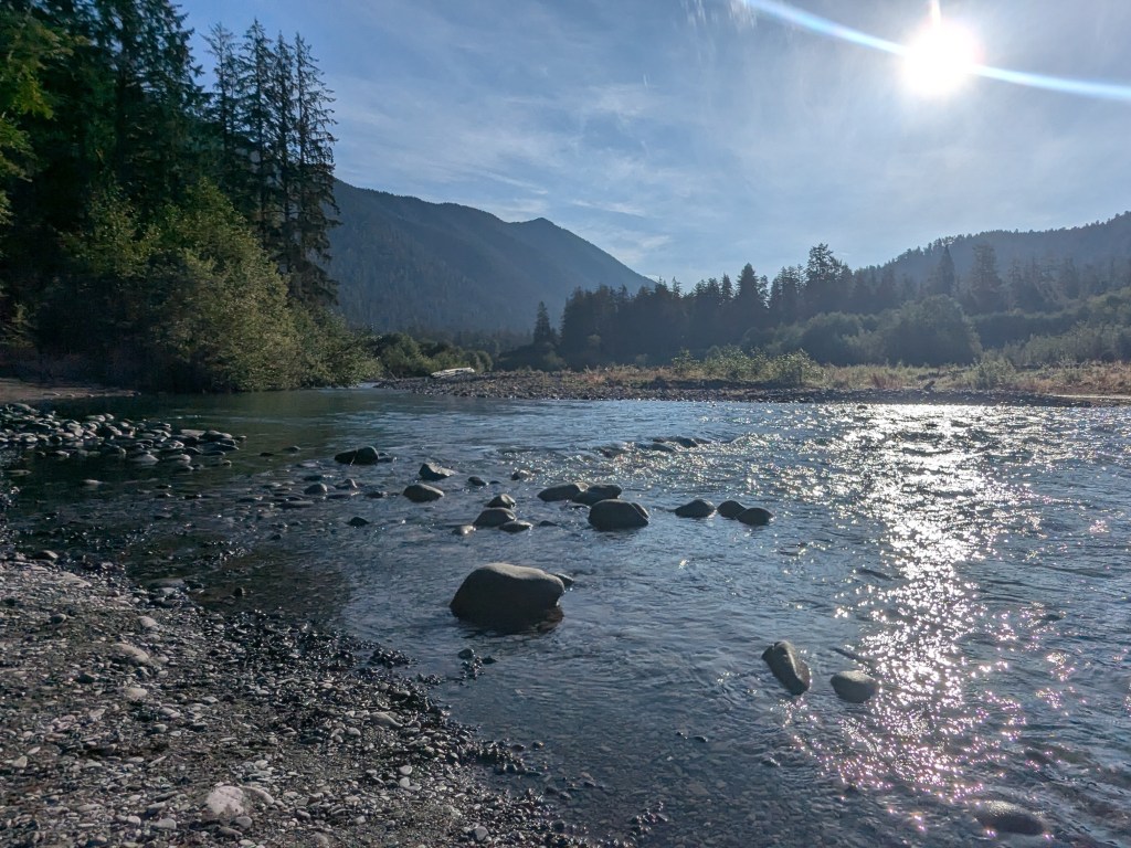 Sun shines on a calm river flowing around large rocks, with mountains and evergreen trees in the background.